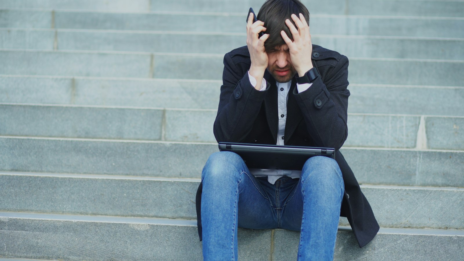 A man sitting on steps with his head in his hands and a laptop on his knees